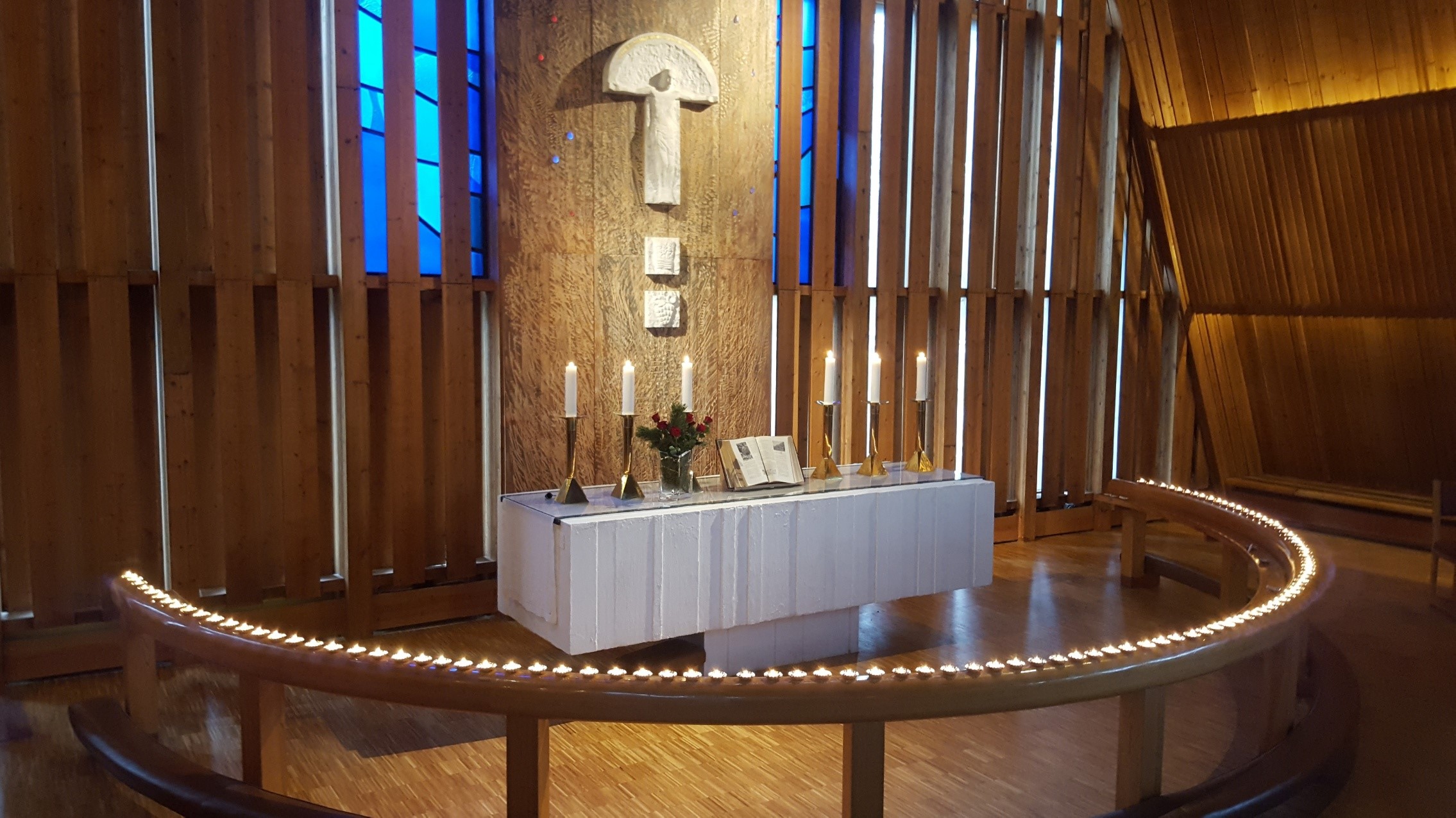 Alterområdet i Brumunddal kirke fremstår med et innbydende og rolig preg. En buet benk omkranset av tente lys fører øyet mot alteret, hvor flere tente stearinlys står langsmed. Veggen bak alteret er dekorert med et kors og geometriske figurer, alt innrammet av høye, vertikale trepaneler som strekker seg opp mot taket. Bakgrunnen er beroligende med en blanding av varmt treverk og diskret belysning som gir et innbydende miljø for ettertanke og ro. Foto.