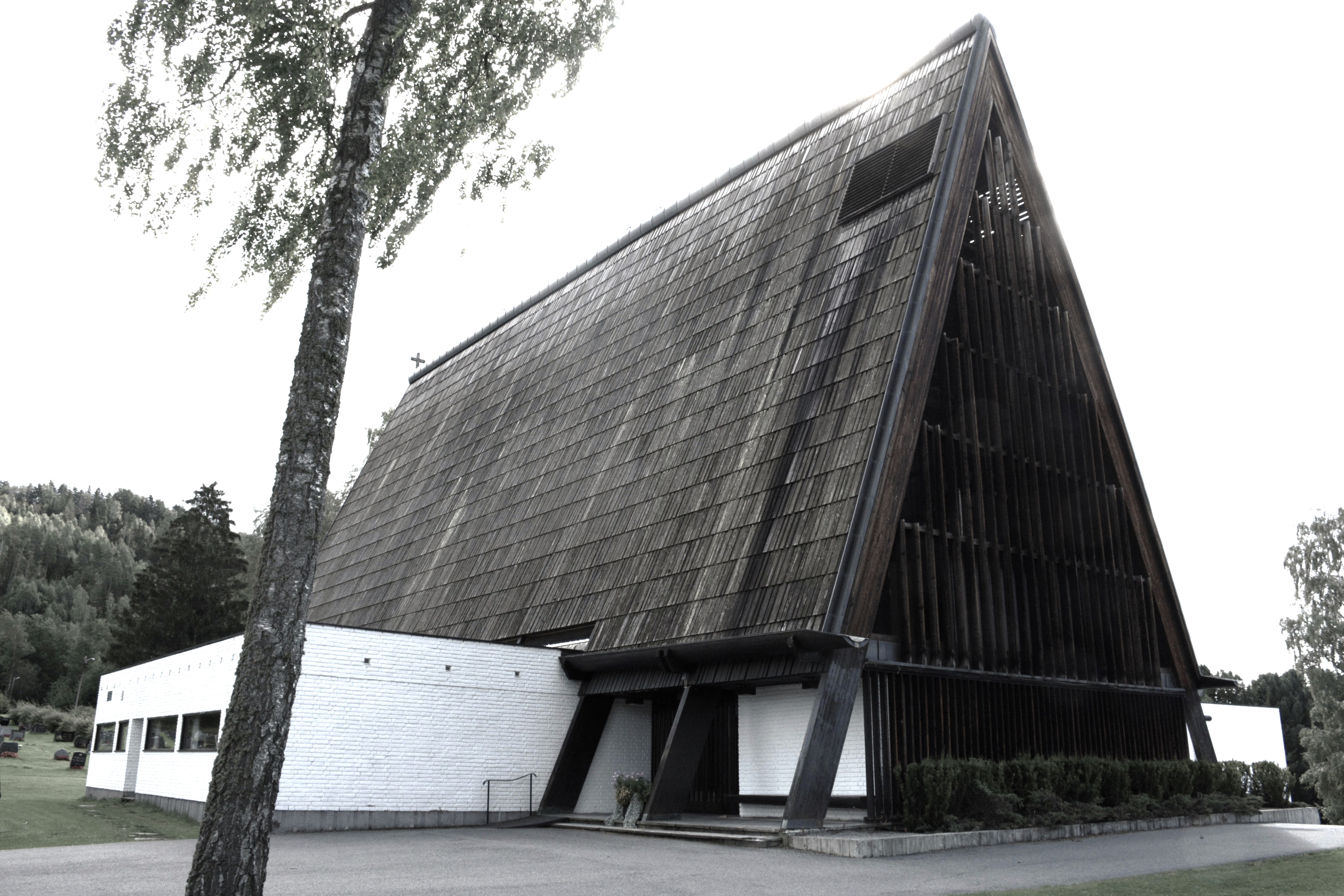 kirke vises her med sin markante arkitektur, preget av et stort, skrånende tak og en struktur av tre og hvit murstein. Kirken står omgitt av grønne trær og en velholdt plen, med en bakgrunn av tett skog som gir en rolig og naturskjønn setting. Arkitekturen er moderne med store, synlige bjelker og en unik form som fanger øyet. Foto.