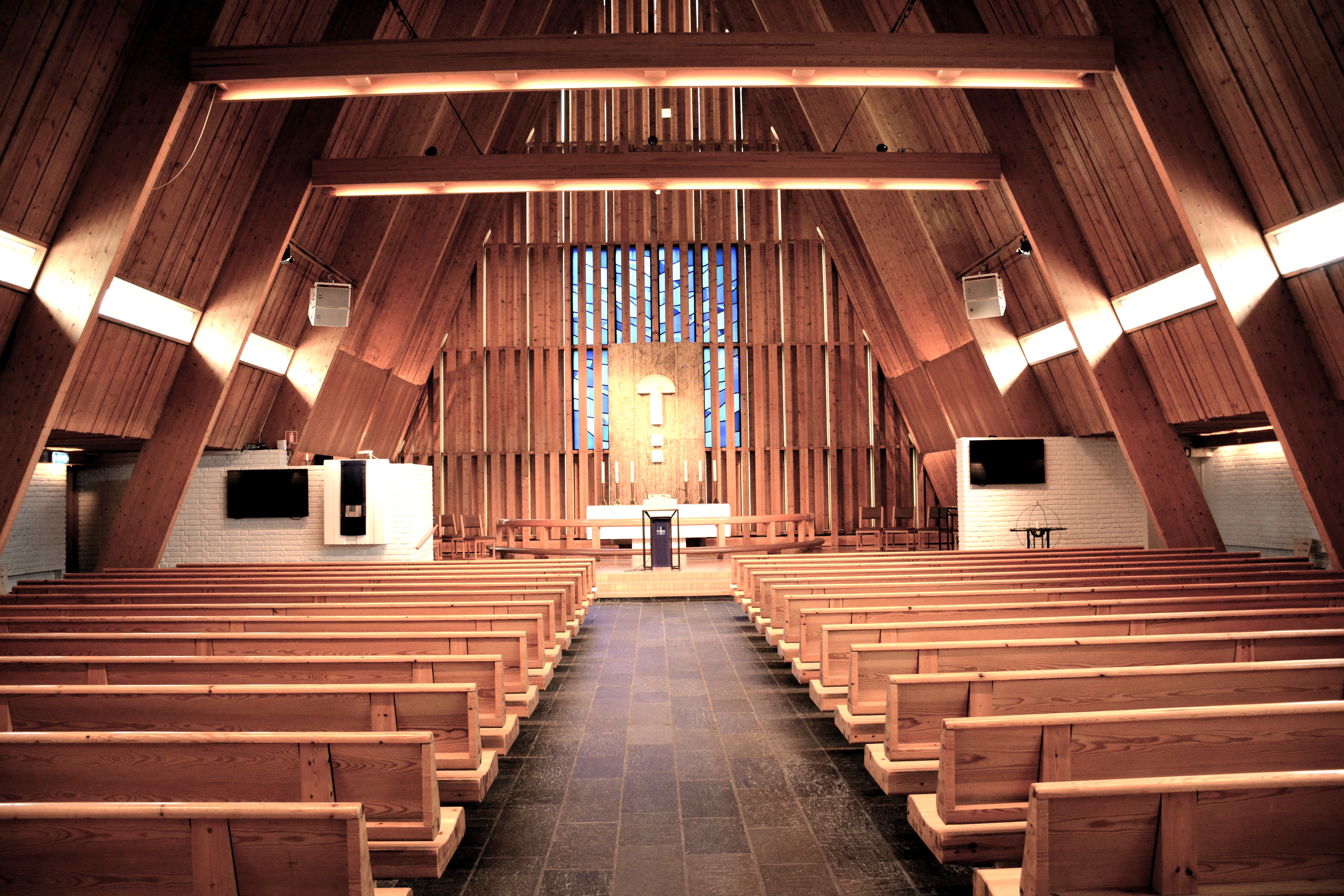 Kirkerommet i Brumunddal kirke er preget av sitt høye, trekledde tak som skråner dramatisk ned mot store vindusflater fylt av farget glass. Den lange midtgangen leder opp til alteret, omgitt av symmetrisk plasserte benkerader. Innredningen er i varmt treverk som harmonerer med den lyse murstrukturen, og skaper et lyst og åpent rom som innbyr til samling og refleksjon. Foto.