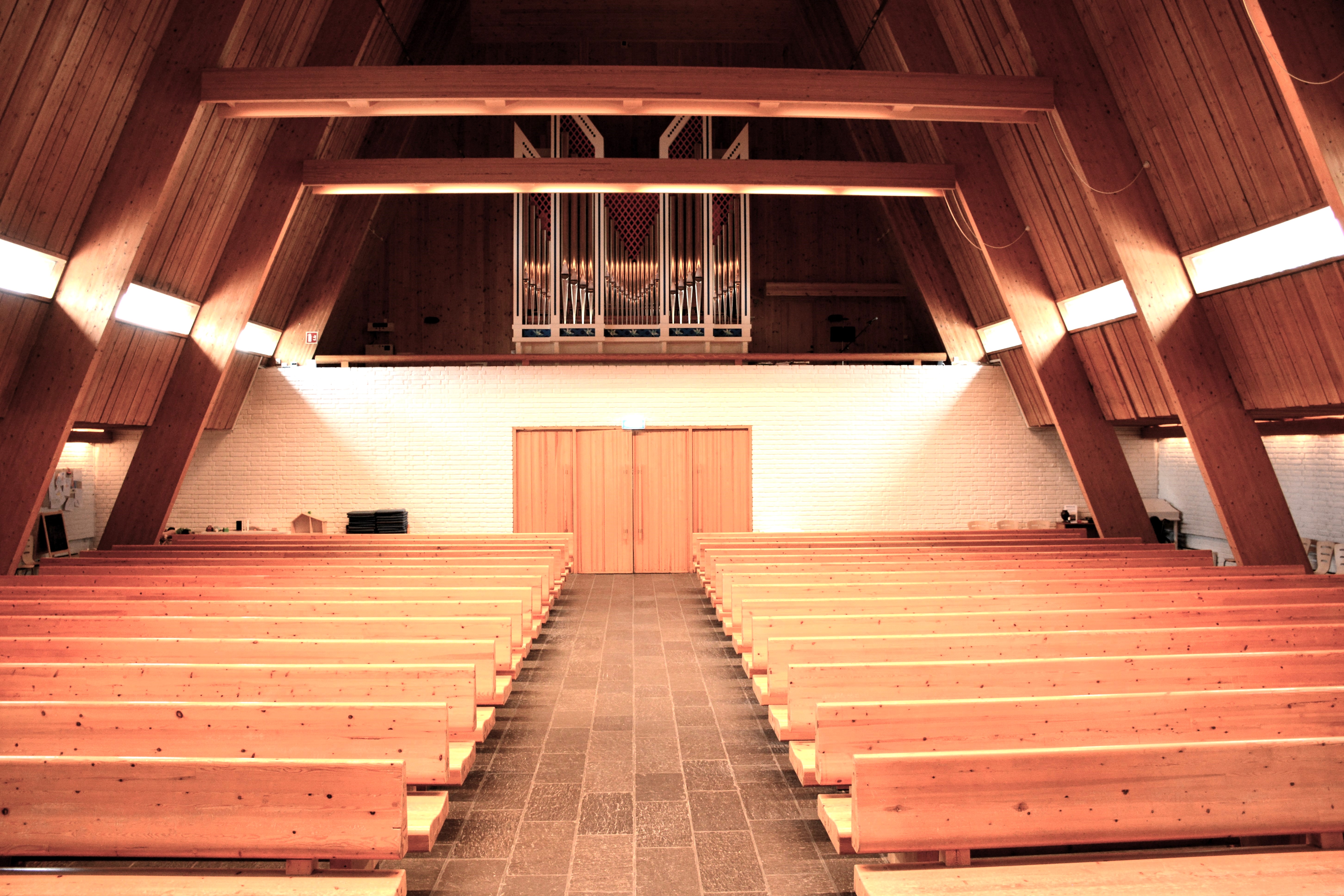 I dette bildet fra Brumunddal kirke ser vi midtgangen som fører opp til orgelområdet, flankert av rekker med trebenker. Orgelpiper dominerer bakveggen, arrangert i et symmetrisk mønster som fanger øyet. Taket og vegger i lyst treverk forsterker rommets akustikk, ideelt for musikalske fremføringer. Denne oppstillingen understreker kirkens arkitektoniske og musikalske arv. Foto.