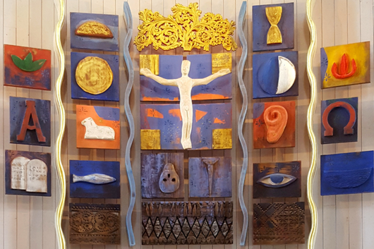 Kunstverket på alteret i Veldre kirke er en detaljert og fargerik sammensetning av ulike religiøse symboler og figurer. Verket består av flere paneler som hver inneholder symboler som et kors, en due, et øye, og andre elementer, alle innrammet av gyldne detaljer. Fargene er livlige og varierte, fra dype blåtoner til glødende rødt og gult, noe som skaper et fokuspunkt som både er åndelig og kunstnerisk tiltalende. Foto.