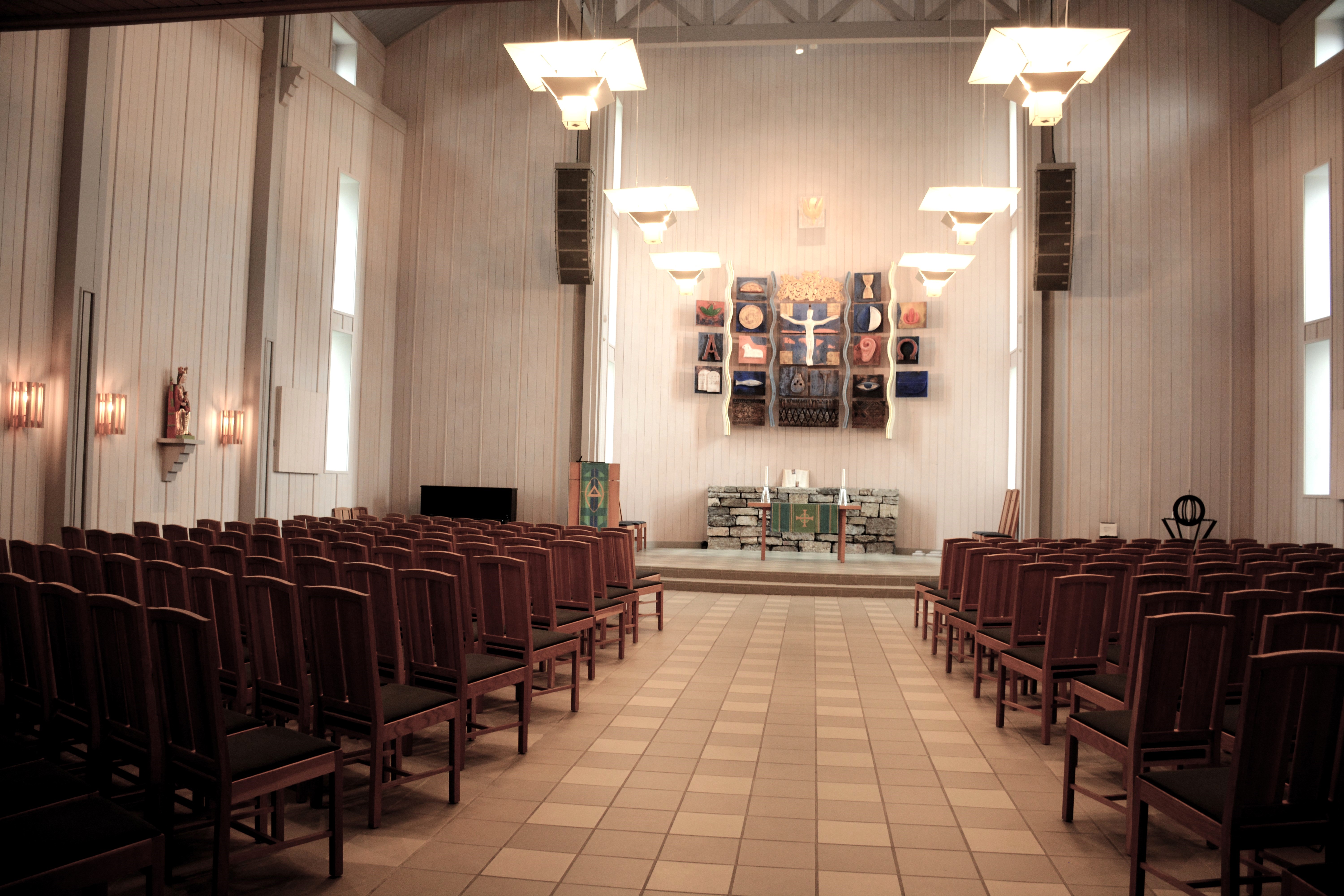 Kirkerommet i Veldre kirke viser en bred midtgang som leder opp til alteret, omringet av rekkevis med brune stoler. Hele rommet er innrammet av lyst treverk og naturlig lys som strømmer inn gjennom vinduene. Den enkle, men elegante innredningen fremhever alteret og kunsten som fungerer som et fokuspunkt for hele rommet. Foto.