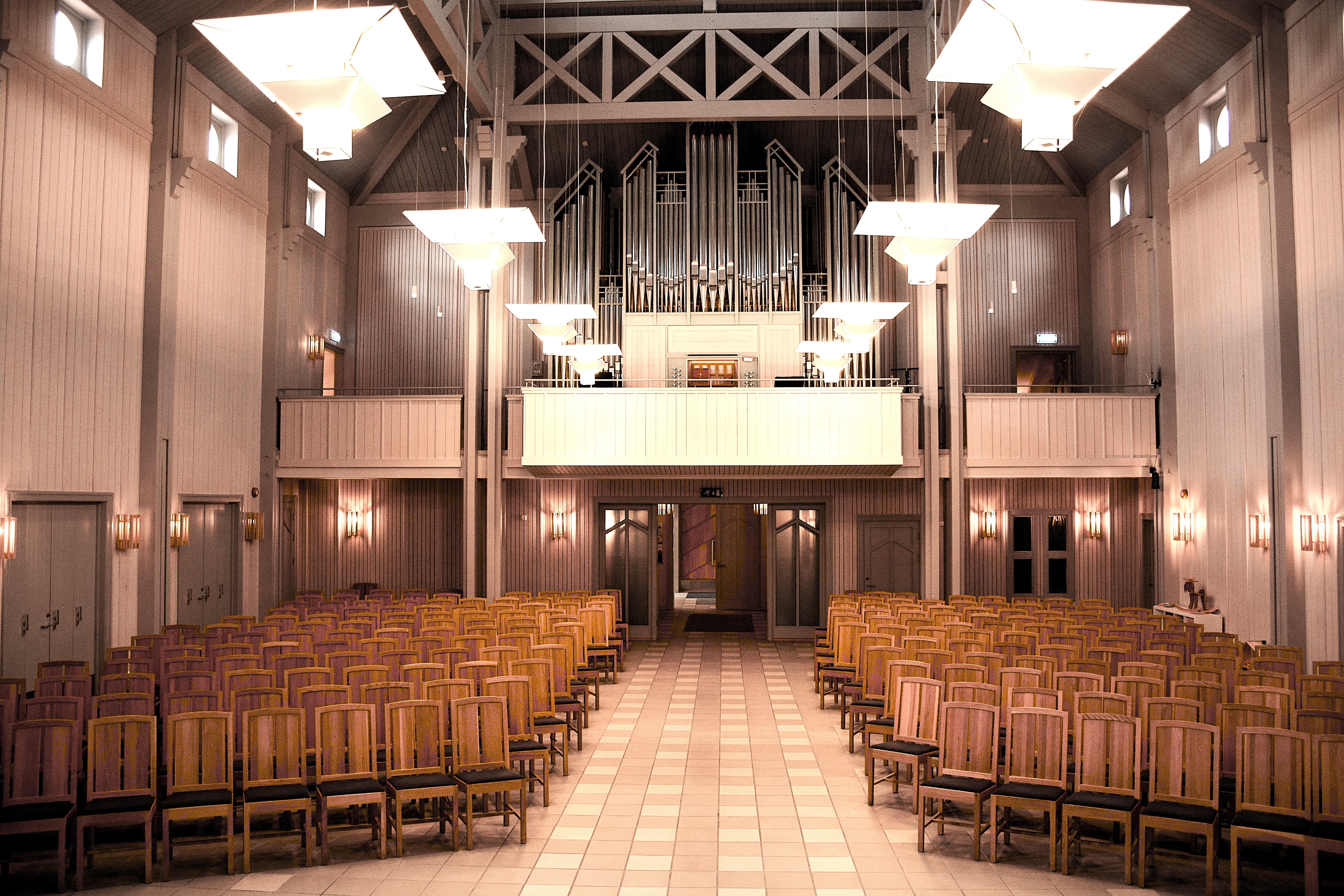 I dette interiøret av Veldre kirke ser vi midtgangen som leder opp til et storslått orgel. Orgelpipene dominerer veggen og skaper en majestetisk bakgrunn for kirkerommet. Rekkene med brune stoler tilfører en følelse av verdighet og tradisjon. Den luftige og åpne arkitekturen styrker akustikken og inviterer til musikalske opplevelser. Foto.