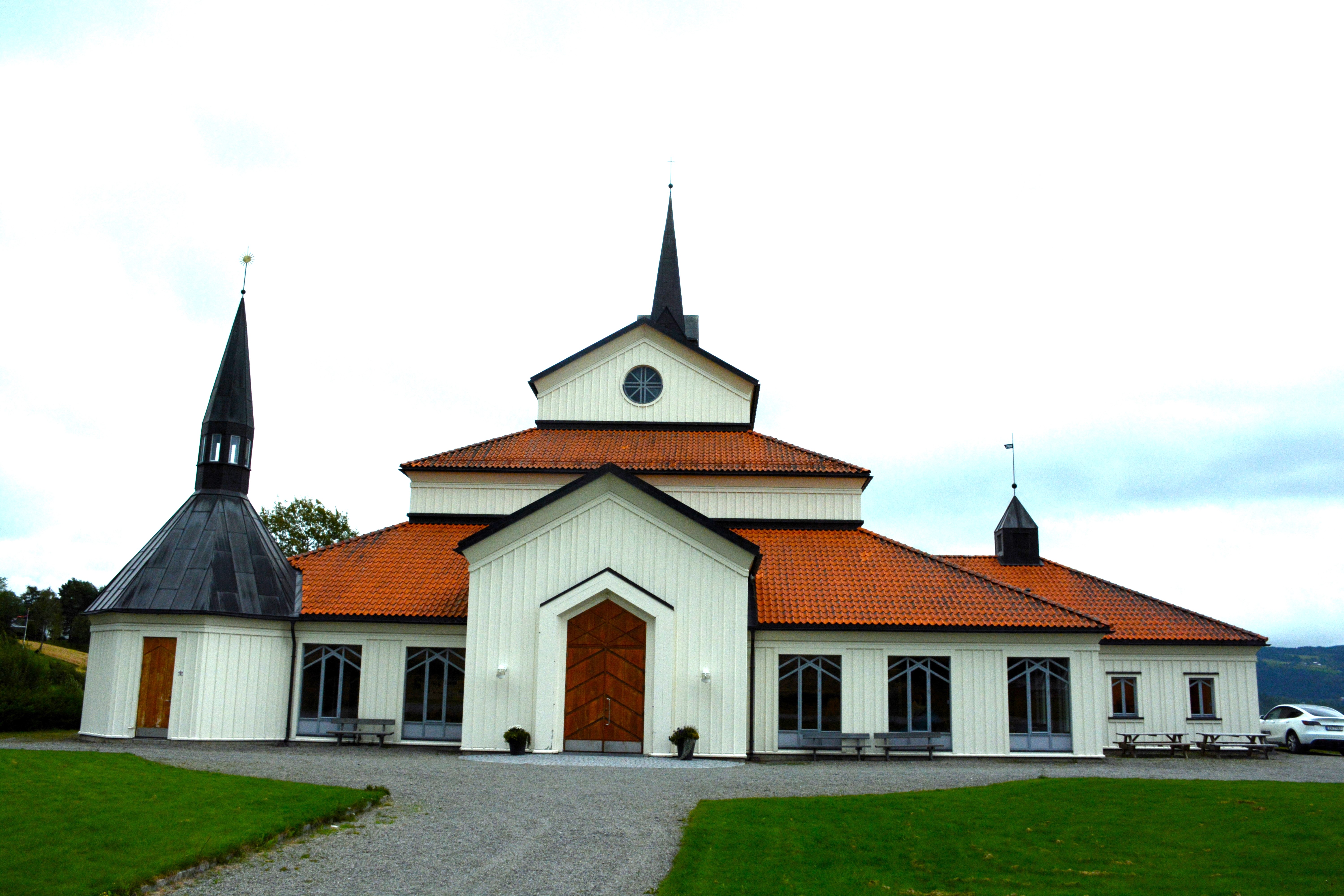 Veldre kirke presenterer en moderne estetikk med sine rene linjer og symmetriske design. Utsiden av kirken er preget av hvite vegger og et stort, spisst tak. Omkringliggende grønne områder og en åpen plass inviterer til samling og fellesskap før og etter kirkelige seremonier. Foto.