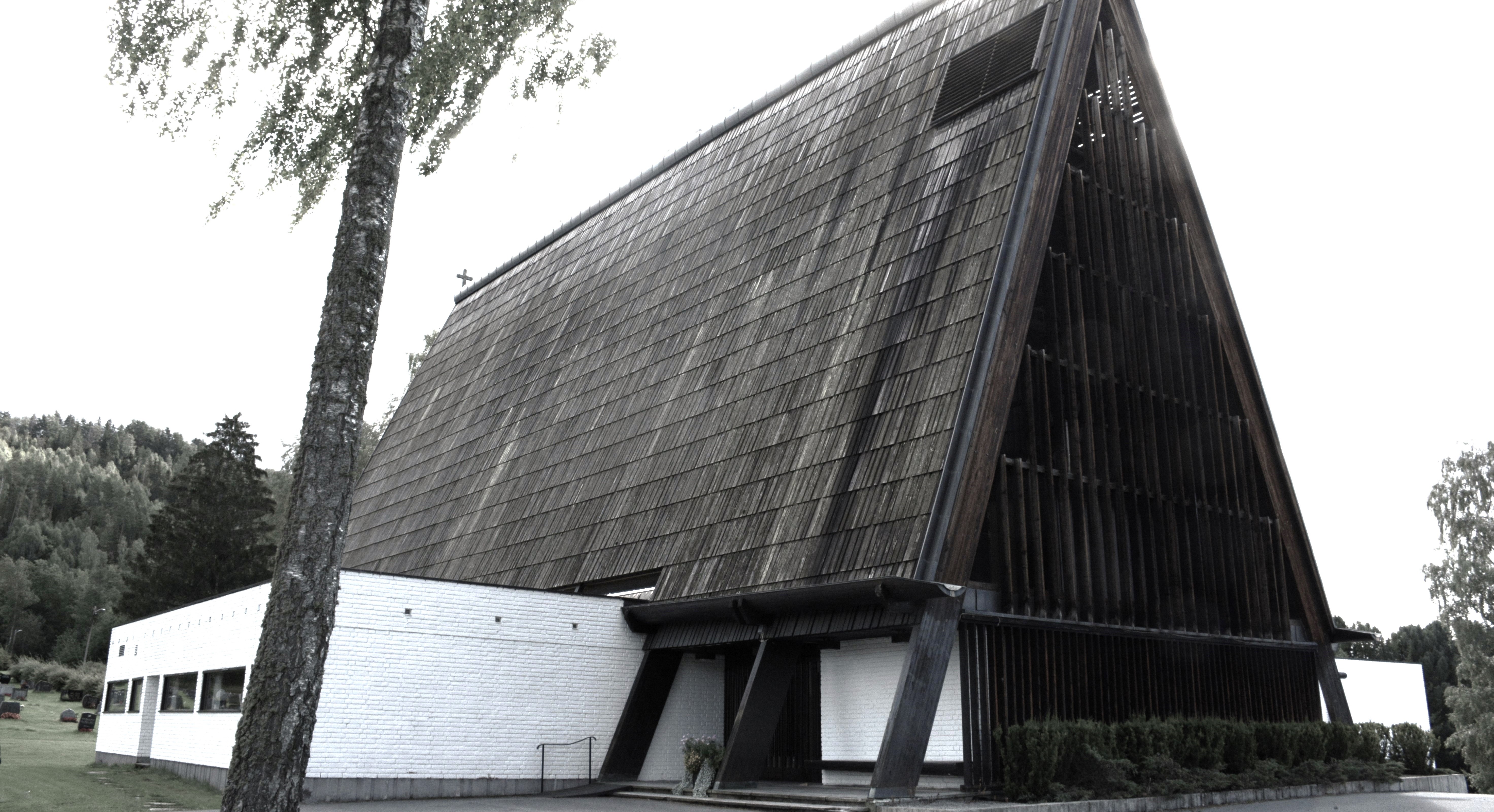 Brumunddal kirke vises her med sin markante arkitektur, preget av et stort, skrånende tak og en struktur av tre og hvit murstein. Kirken står omgitt av grønne trær og en velholdt plen, med en bakgrunn av tett skog som gir en rolig og naturskjønn setting. Arkitekturen er moderne med store, synlige bjelker og en unik form som fanger øyet. Foto.