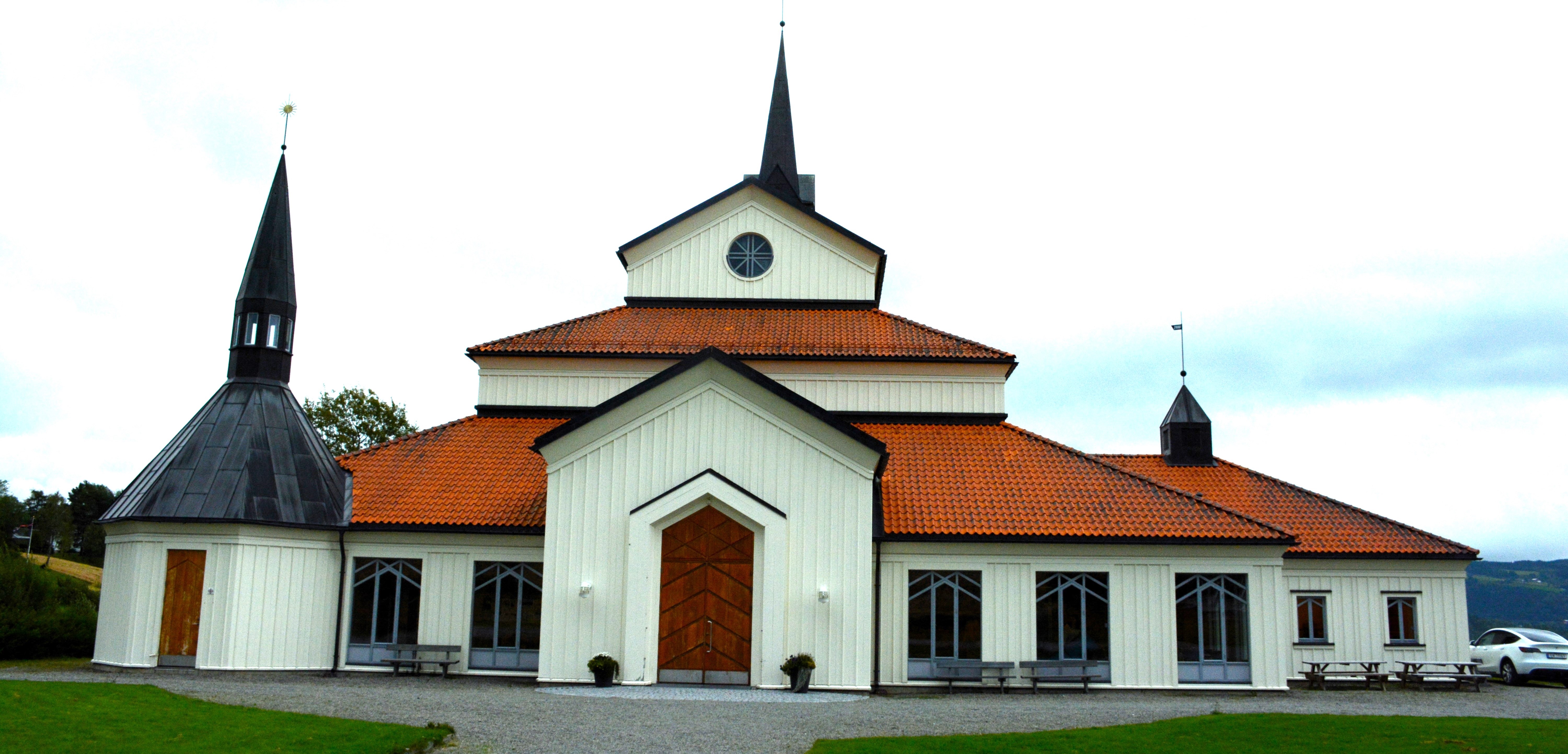 Veldre kirke presenterer en moderne estetikk med sine rene linjer og symmetriske design. Utsiden av kirken er preget av hvite vegger og et stort, spisst tak. Omkringliggende grønne områder og en åpen plass inviterer til samling og fellesskap før og etter kirkelige seremonier. Foto.